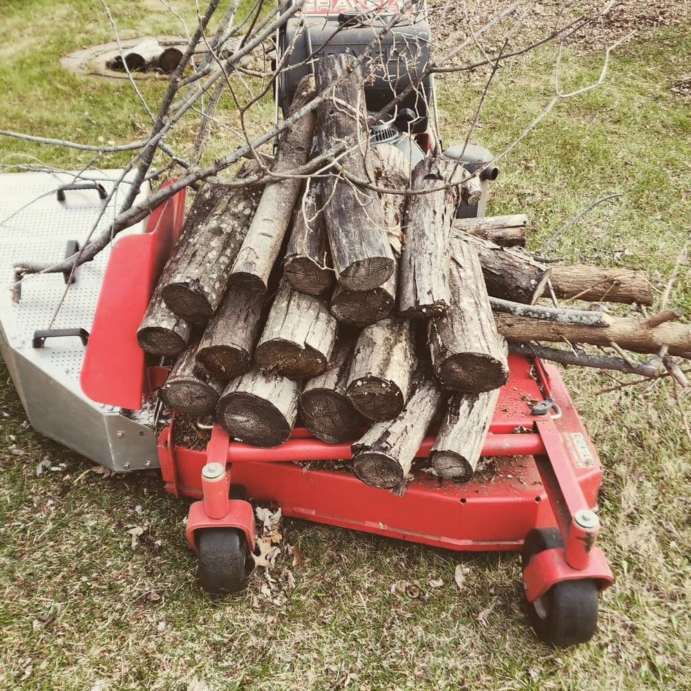 Red lawn mower stacked with firewood logs and branches in a grassy yard setting.